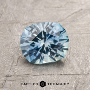 A 1.25-carat Sky Blue Montana Sapphire (heated) with a starburst cut is shown on a textured gray surface, featuring the "Earth's Treasury" logo at the bottom.