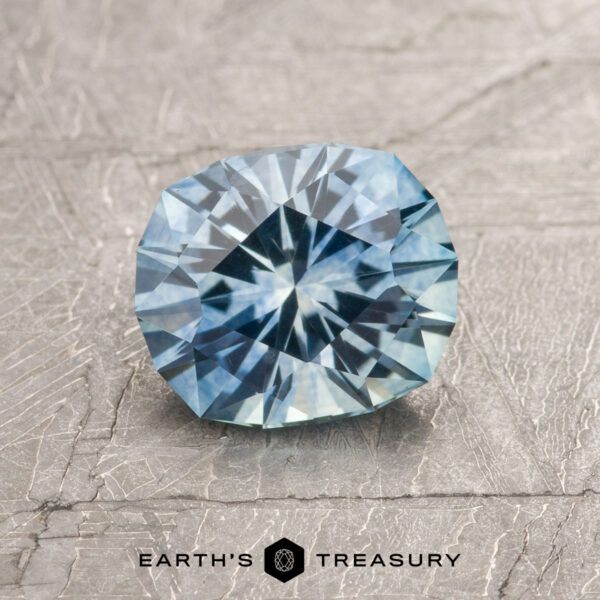 1.25-Carat Sky Blue Montana Sapphire (Heated) A 1.25-carat Sky Blue Montana Sapphire (heated) with a starburst cut is shown on a textured gray surface, featuring the "Earth's Treasury" logo at the bottom.