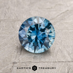 A 1.43-carat heated teal blue Montana sapphire is displayed on a textured light gray surface, with the "Earth's Treasury" logo and text at the bottom.