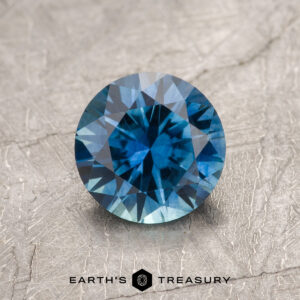 A 1.41-carat Peacock Blue Montana Sapphire (Heated), round-cut with multiple facets, is displayed on a textured surface. The text "EARTH'S TREASURY" appears at the bottom.