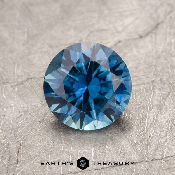 1.41-Carat Peacock Blue Montana Sapphire (Heated) A 1.41-carat Peacock Blue Montana Sapphire (Heated), round-cut with multiple facets, is displayed on a textured surface. The text "EARTH'S TREASURY" appears at the bottom.