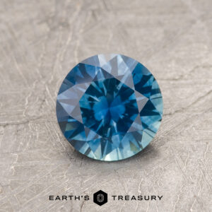 1.29-Carat Peacock Blue Montana Sapphire (Heated), round brilliant-cut, displayed on a textured gray surface with "EARTH'S TREASURY" below the gem.
