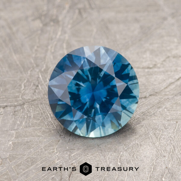 1.29-Carat Peacock Blue Montana Sapphire (Heated) 1.29-Carat Peacock Blue Montana Sapphire (Heated), round brilliant-cut, displayed on a textured gray surface with "EARTH'S TREASURY" below the gem.