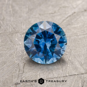 A 1.29-carat medium blue Montana sapphire (heated) is displayed on a textured gray surface, with the text "EARTH'S TREASURY" at the bottom.