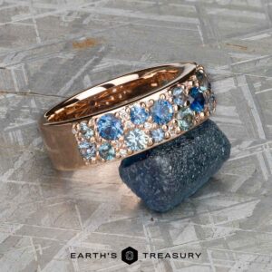 The Thin "Borealis" ring in 14k rose gold, featuring teal sapphire and white gemstones, rests on a rough blue crystal atop a textured surface, with "EARTH'S TREASURY" below.
