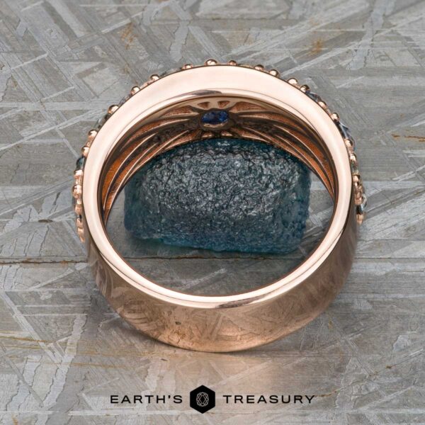 The Thin "Borealis" ring in 14k rose gold The Thin "Borealis" ring in 14k rose gold, featuring intricate detailing and a teal sapphire gemstone, is displayed upright on a textured gray surface above the Earth's Treasury logo.