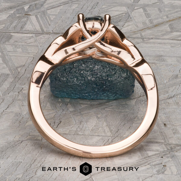 The "Aislinn" ring in 14k rose gold with 1.69-carat Montana sapphire The "Aislinn" ring in 14k rose gold features a 1.69-carat Montana sapphire and an intricate band, standing upright on a textured grey background with the Earth's Treasury logo at the bottom.