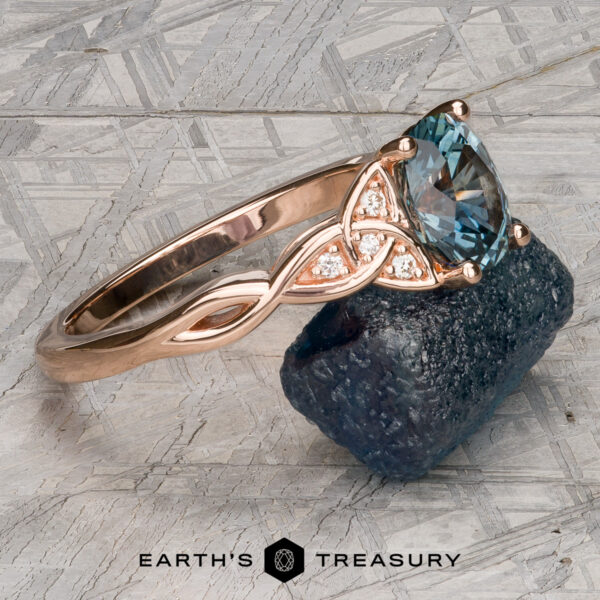 The "Aislinn" ring in 14k rose gold with 1.69-carat Montana sapphire The "Aislinn" ring in 14k rose gold, featuring a 1.69-carat Montana sapphire and small diamonds, is displayed atop a dark mineral on a textured gray surface, with the "Earth's Treasury" logo below.