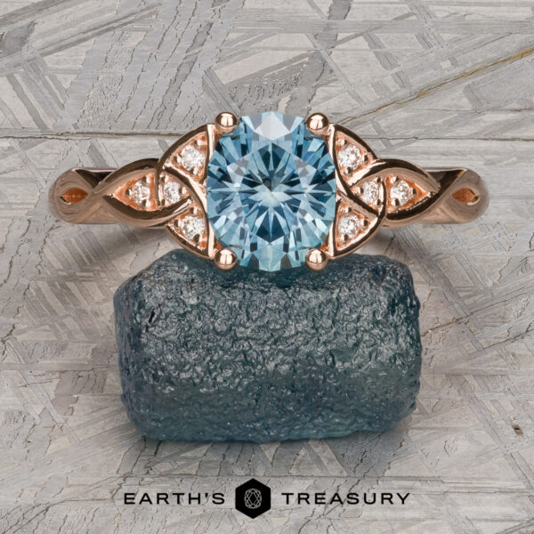The "Aislinn" ring in 14k rose gold with 1.69-carat Montana sapphire Stacked from 53 images (Method=B, R=1, S=3), the rose gold Aislinn Ring with an oval aquamarine center and diamond accents is shown on a rough blue mineral, set against a geometric background.