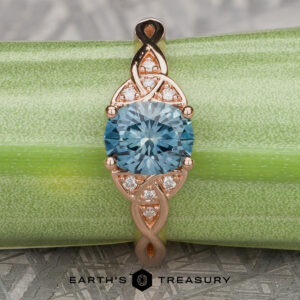 The “Aislinn” ring in 14k rose gold showcases a 1.69-carat Montana sapphire and Celtic-inspired details, elegantly photographed on a green textured background.