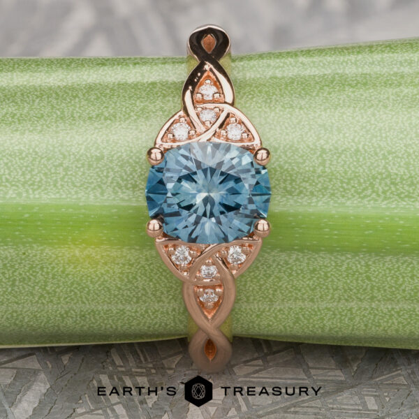 The “Aislinn” ring in 14k rose gold showcases a 1.69-carat Montana sapphire and Celtic-inspired details, elegantly photographed on a green textured background.