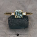 A "Finella" ring in 18K yellow gold with a 0.95-carat light green Montana sapphire and two rectangular side stones, displayed on a dark textured rock. The "Earth's Treasury" logo appears at the bottom.