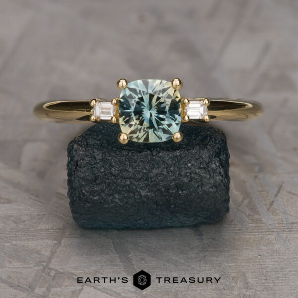 The "Finella" ring in 18K yellow gold with 0.95-Carat Montana sapphire A "Finella" ring in 18K yellow gold with a 0.95-carat light green Montana sapphire and two rectangular side stones, displayed on a dark textured rock. The "Earth's Treasury" logo appears at the bottom.