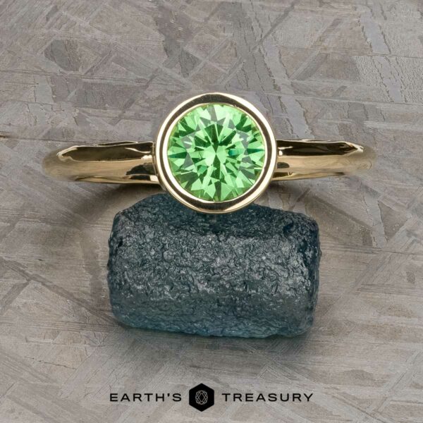 The "Emma" ring in 14k yellow gold with 1.49-carat Tsavorite Garnet, wave texture band The "Emma" ring in 14k yellow gold with a 1.49-carat Tsavorite Garnet and wave texture band rests on a rough, dark blue stone with the text "EARTH'S TREASURY" below.
