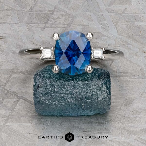 The "Finella" ring in platinum with 2.13-carat Montana sapphire The "Finella" ring in platinum features a 2.13-carat Montana sapphire center stone flanked by two clear stones, displayed on rough blue crystal with the "Earth's Treasury" logo below.
