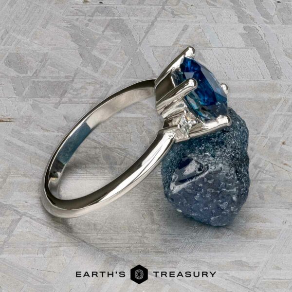 The "Finella" ring in platinum with 2.13-carat Montana sapphire The "Finella" platinum ring with a 2.13-carat Montana sapphire rests on a matching rough blue stone atop a textured gray surface, with the "Earth's Treasury" logo displayed at the bottom.