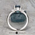 The "Finella" ring in platinum with a 2.13-carat Montana sapphire is shown upright on a textured gray stone background, sparkling side accents aglow. "EARTH'S TREASURY" appears at the bottom.