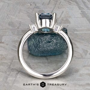 The "Finella" ring in platinum with a 2.13-carat Montana sapphire is shown upright on a textured gray stone background, sparkling side accents aglow. "EARTH'S TREASURY" appears at the bottom.