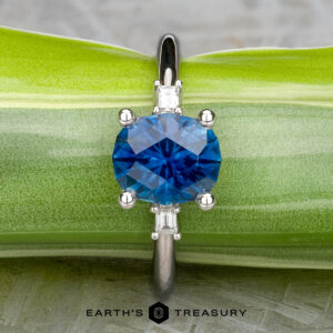 The "Finella" ring in platinum, featuring a 2.13-carat Montana sapphire and small clear side stones, is displayed on a green leaf with the "EARTH'S TREASURY" logo at the bottom.