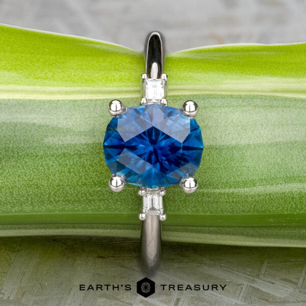 The "Finella" ring in platinum with 2.13-carat Montana sapphire The "Finella" ring in platinum, featuring a 2.13-carat Montana sapphire and small clear side stones, is displayed on a green leaf with the "EARTH'S TREASURY" logo at the bottom.
