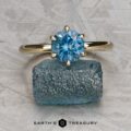 The Minimal "Anemone" ring in 14k yellow gold with a 1.46-carat Montana sapphire is showcased solitaire atop a rough blue crystal, against a textured gray background. The text "EARTH'S TREASURY" appears below.