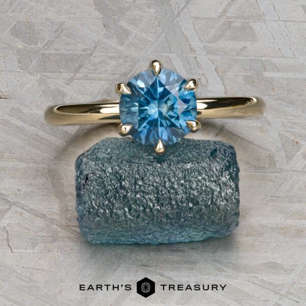 The Minimal "Anemone" ring in 14k yellow gold with 1.46-carat Montana sapphire The Minimal "Anemone" ring in 14k yellow gold with a 1.46-carat Montana sapphire is showcased solitaire atop a rough blue crystal, against a textured gray background. The text "EARTH'S TREASURY" appears below.