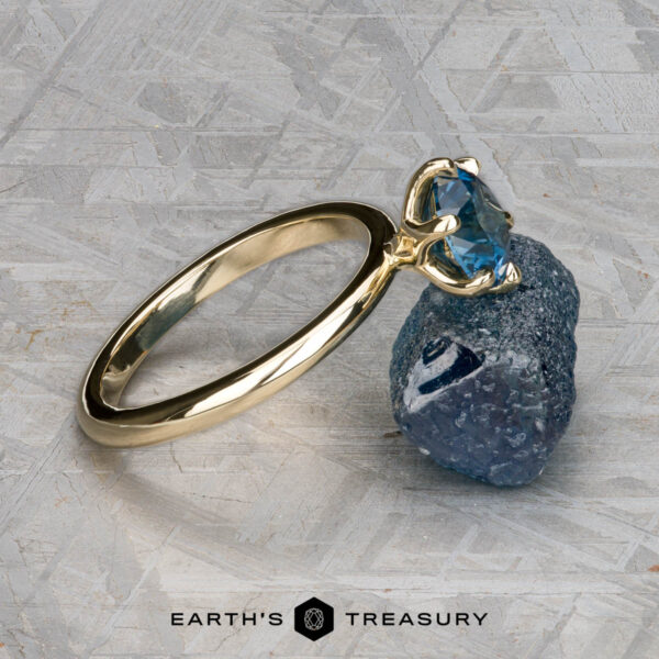 The Minimal "Anemone" ring in 14K yellow gold with 1.46-carat Montana sapphire The Minimal "Anemone" ring in 14k yellow gold with a 1.46-carat Montana sapphire is showcased solitaire atop a rough blue crystal, against a textured gray background. The text "EARTH'S TREASURY" appears below.
