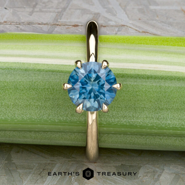 The Minimal "Anemone" ring in 14K yellow gold with 1.46-carat Montana sapphire The Minimal "Anemone" ring in 14K yellow gold features a 1.46-carat round Montana sapphire in a six-prong solitaire setting, shown on a green background with "Earth's Treasury" below.