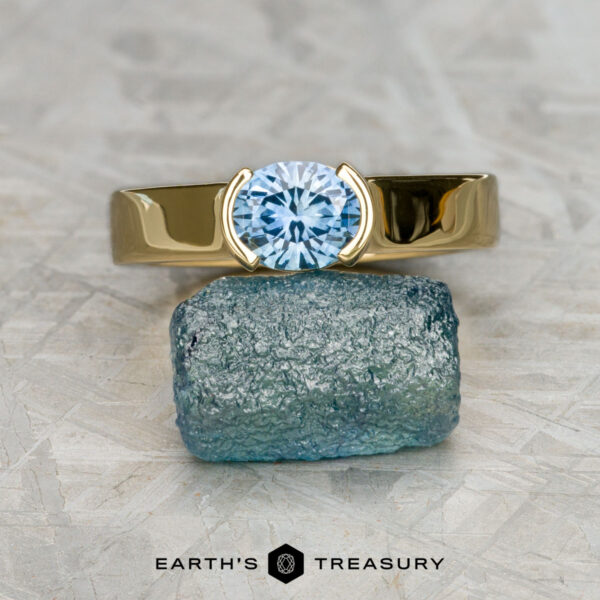 The Wide "Larisa" ring in 18K yellow gold with 1.13-carat Montana sapphire The Wide "Larisa" ring in 18K yellow gold with a 1.13-carat Montana sapphire is shown balanced on a rough blue-green stone. "EARTH'S TREASURY" is displayed at the bottom of the image.