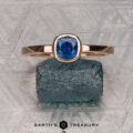 A 0.76-carat Montana sapphire Low-Profile "Emma" ring in 14K rose gold rests on a rough blue stone atop a patterned metallic surface. Below, "EARTH'S TREASURY" highlights the ring’s elegant solitaire design.