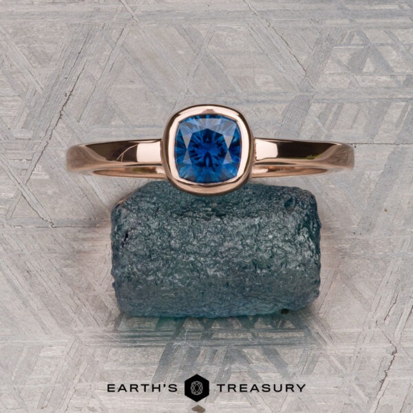 The Low-Profile "Emma" ring in 14K rose gold with 0.76-carat Montana sapphire A 0.76-carat Montana sapphire Low-Profile "Emma" ring in 14K rose gold rests on a rough blue stone atop a patterned metallic surface. Below, "EARTH'S TREASURY" highlights the ring’s elegant solitaire design.