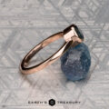 A 0.76-carat Montana sapphire Low-Profile "Emma" ring in 14K rose gold rests on a rough blue stone atop a patterned metallic surface. Below, "EARTH'S TREASURY" highlights the ring’s elegant solitaire design.