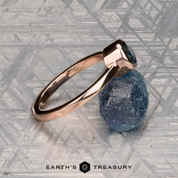 The Low-Profile "Emma" ring in 14K rose gold with 0.76-carat Montana sapphire A 0.76-carat Montana sapphire Low-Profile "Emma" ring in 14K rose gold rests on a rough blue stone atop a patterned metallic surface. Below, "EARTH'S TREASURY" highlights the ring’s elegant solitaire design.