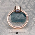 A 0.76-carat Montana sapphire Low-Profile "Emma" ring in 14K rose gold rests on a rough blue stone atop a patterned metallic surface. Below, "EARTH'S TREASURY" highlights the ring’s elegant solitaire design.