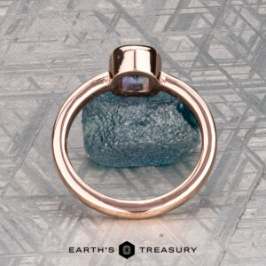 A 0.76-carat Montana sapphire Low-Profile "Emma" ring in 14K rose gold rests on a rough blue stone atop a patterned metallic surface. Below, "EARTH'S TREASURY" highlights the ring’s elegant solitaire design.