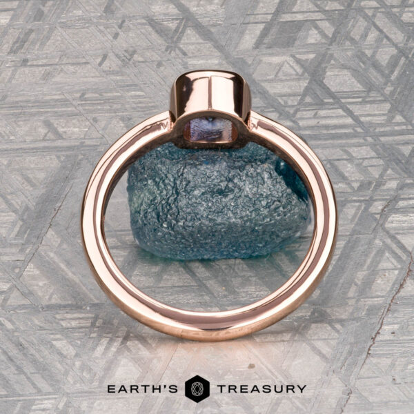 The Low-Profile "Emma" ring in 14K rose gold with 0.76-carat Montana sapphire A 0.76-carat Montana sapphire Low-Profile "Emma" ring in 14K rose gold rests on a rough blue stone atop a patterned metallic surface. Below, "EARTH'S TREASURY" highlights the ring’s elegant solitaire design.