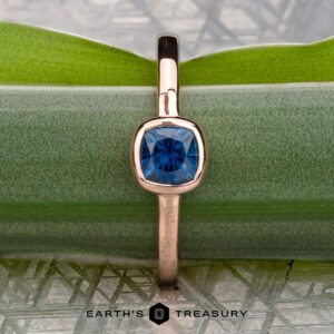 The Low-Profile "Emma" ring in 14K rose gold with a 0.76-carat Montana sapphire is shown vertically on a green leaf, with "Earth's Treasury" displayed below.