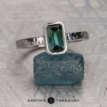 A Vigh, Melissa 27 ring with a rectangular green gemstone sits on a rough blue stone atop a textured gray surface, the "Earth's Treasury" logo beneath.