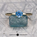 The “Felicia” ring in 14K yellow gold featuring a 0.81-carat Montana sapphire stands upright on a rough blue mineral, with a hidden halo detail, set against a geometric background. The "Earth's Treasury" logo appears at the bottom.