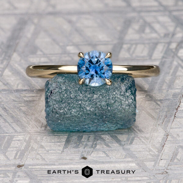 The "Felicia" ring in 14K yellow gold with 0.81-carat Montana sapphire The “Felicia” ring in 14K yellow gold featuring a 0.81-carat Montana sapphire stands upright on a rough blue mineral, with a hidden halo detail, set against a geometric background. The "Earth's Treasury" logo appears at the bottom.