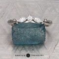 The "Magnolia" band in platinum, featuring three marquise and pear-cut gemstones, rests on a rough blue crystal with a geometric patterned background and the “Earth’s Treasury” logo below.