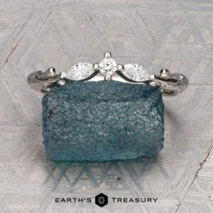 The "Magnolia" band in platinum, featuring three marquise and pear-cut gemstones, rests on a rough blue crystal with a geometric patterned background and the “Earth’s Treasury” logo below.