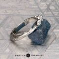 The "Magnolia" band in platinum, featuring three marquise and pear-cut gemstones, rests on a rough blue crystal with a geometric patterned background and the “Earth’s Treasury” logo below.
