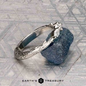 The "Magnolia" band in platinum, featuring three marquise and pear-cut gemstones, rests on a rough blue crystal with a geometric patterned background and the “Earth’s Treasury” logo below.