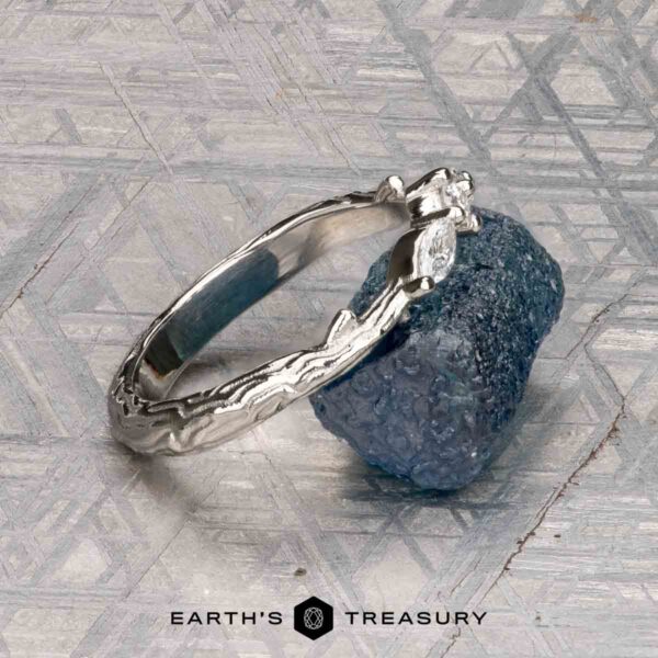 The "Magnolia" band in platinum The "Magnolia" band in platinum, featuring three marquise and pear-cut gemstones, rests on a rough blue crystal with a geometric patterned background and the “Earth’s Treasury” logo below.