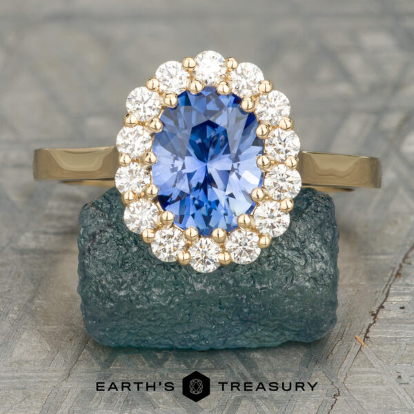 The “Elaine” ring in 18K yellow gold with 1.75-carat Ceylon sapphire The “Elaine” ring in 18K yellow gold features a stunning 1.75-carat Ceylon sapphire framed by round white diamonds, shown upright on textured stone above the "Earth's Treasury" logo—an exquisite engagement ring.
