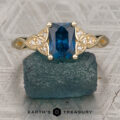 A gold Noel, Jonathan ring with a rectangular blue gemstone and small diamonds is displayed on a rough blue stone. The text "EARTH'S TREASURY" appears at the bottom of the image.