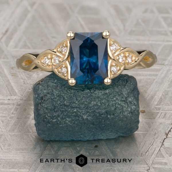 The "Aislinn" Ring A gold ring with a rectangular blue gemstone and small diamonds is displayed on a rough blue stone. The text "EARTH'S TREASURY" appears at the bottom of the image.