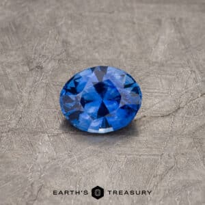 A 2.30-carat Royal Blue Ceylon Sapphire (heated), expertly cut and polished, lies on a textured gray surface. The brand "Earth's Treasury" is visible at the bottom in black text.
