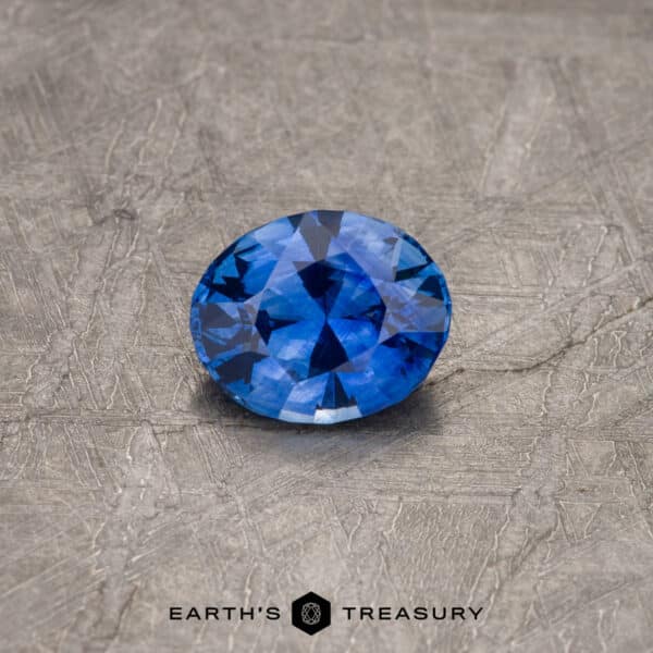 1.56-Carat Ceylon Sapphire (Heated) A 2.30-carat Royal Blue Ceylon Sapphire (heated), expertly cut and polished, lies on a textured gray surface. The brand "Earth's Treasury" is visible at the bottom in black text.